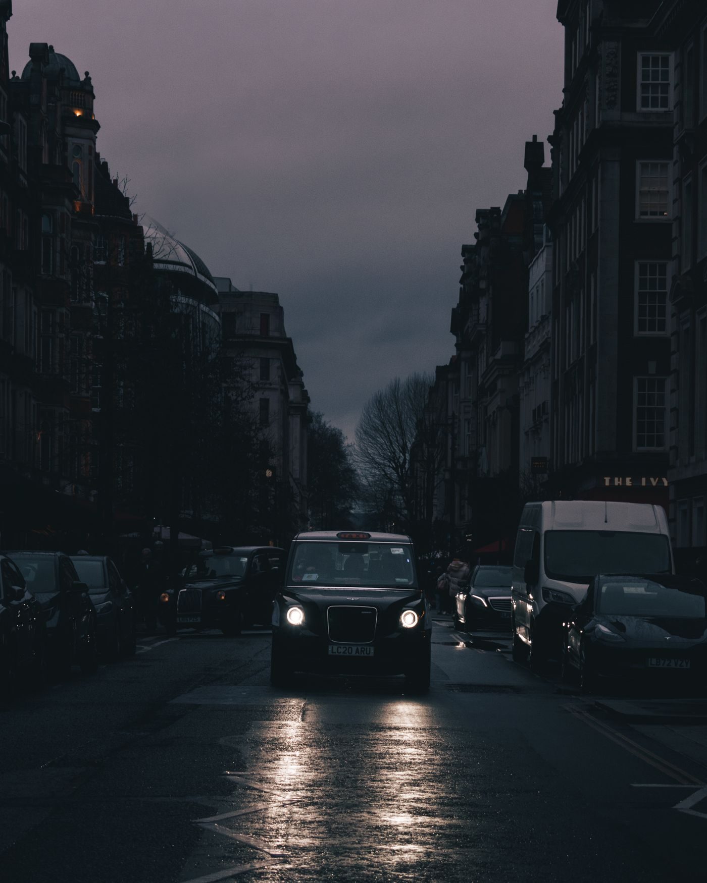 Wet London street at dusk with car headlights and dark mansion blocks either side